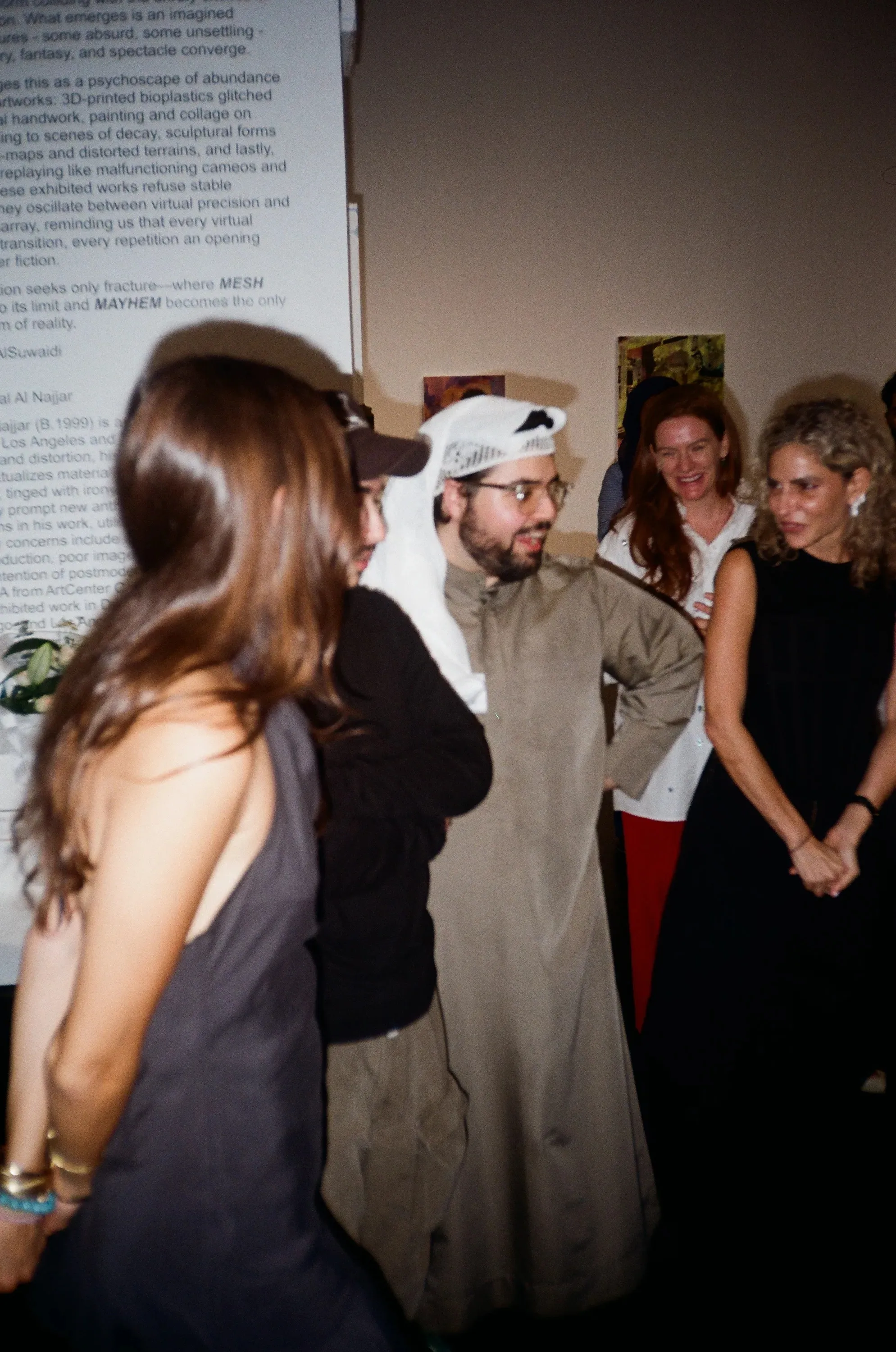 A guest in a keffiyeh laughing with others in front of the exhibition wall text and framed artworks