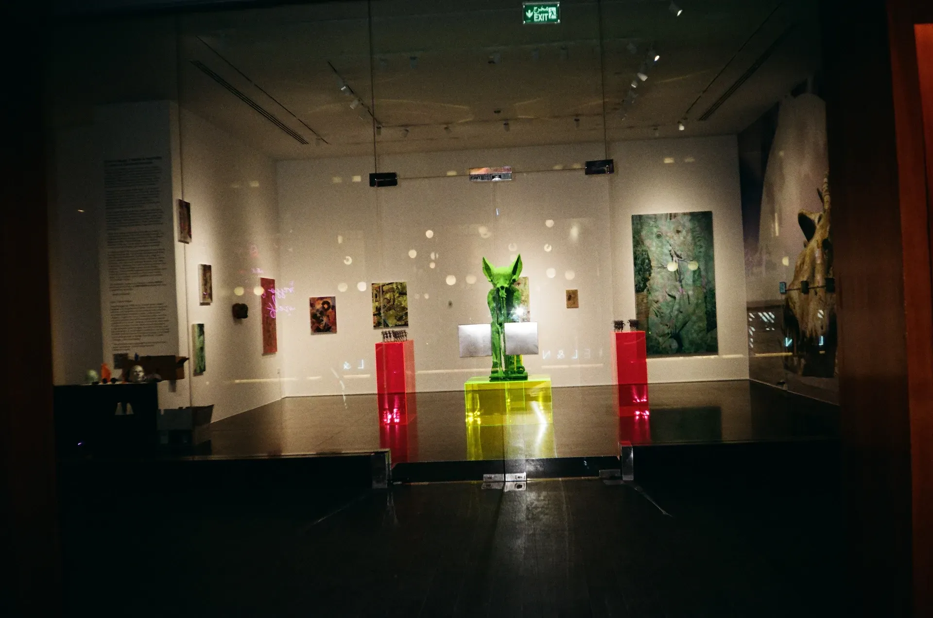 A gallery room with paintings on the walls and a glowing green sculptural figure displayed on illuminated pedestals