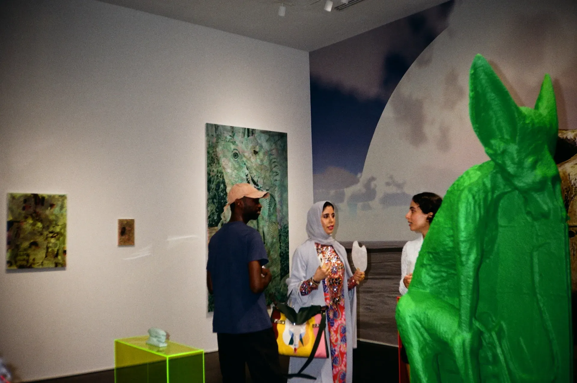 Gallery visitors, including one in a bright green sculptural costume, talking beside paintings and a wall-sized photographic mural