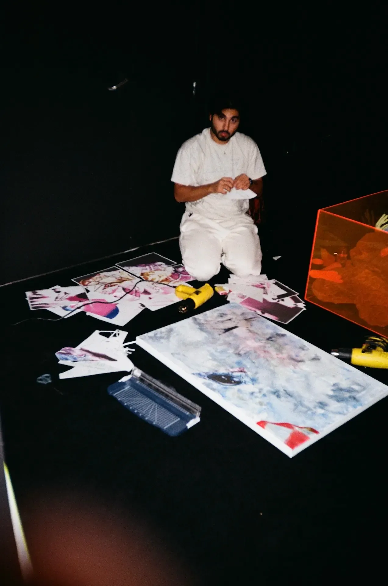 An artist kneeling on a dark studio floor surrounded by printed photographs, a glue gun, and a paint-splattered canvas