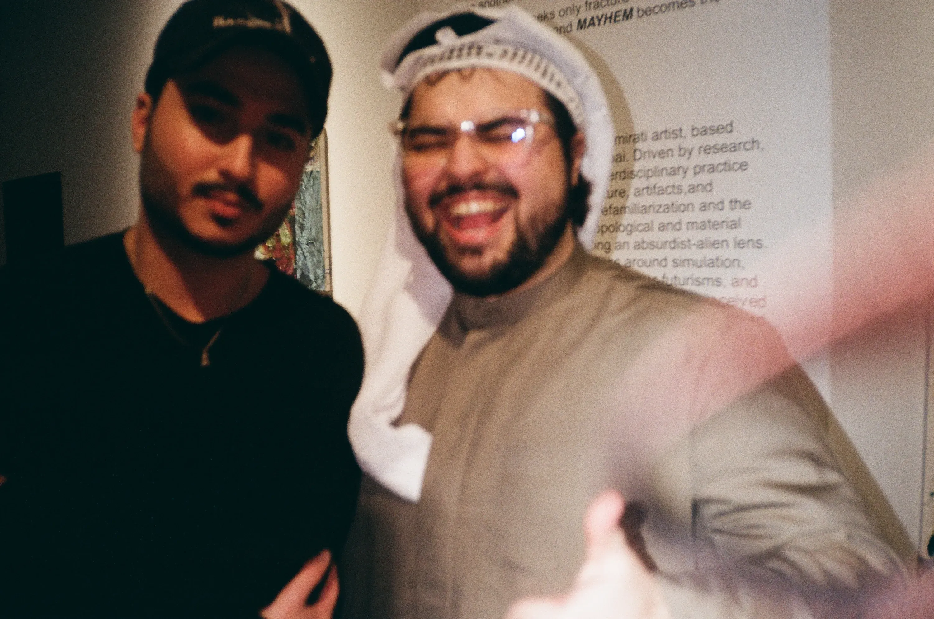 Two guests laughing in front of the exhibition's wall text, one wearing a checkered keffiyeh-style headscarf