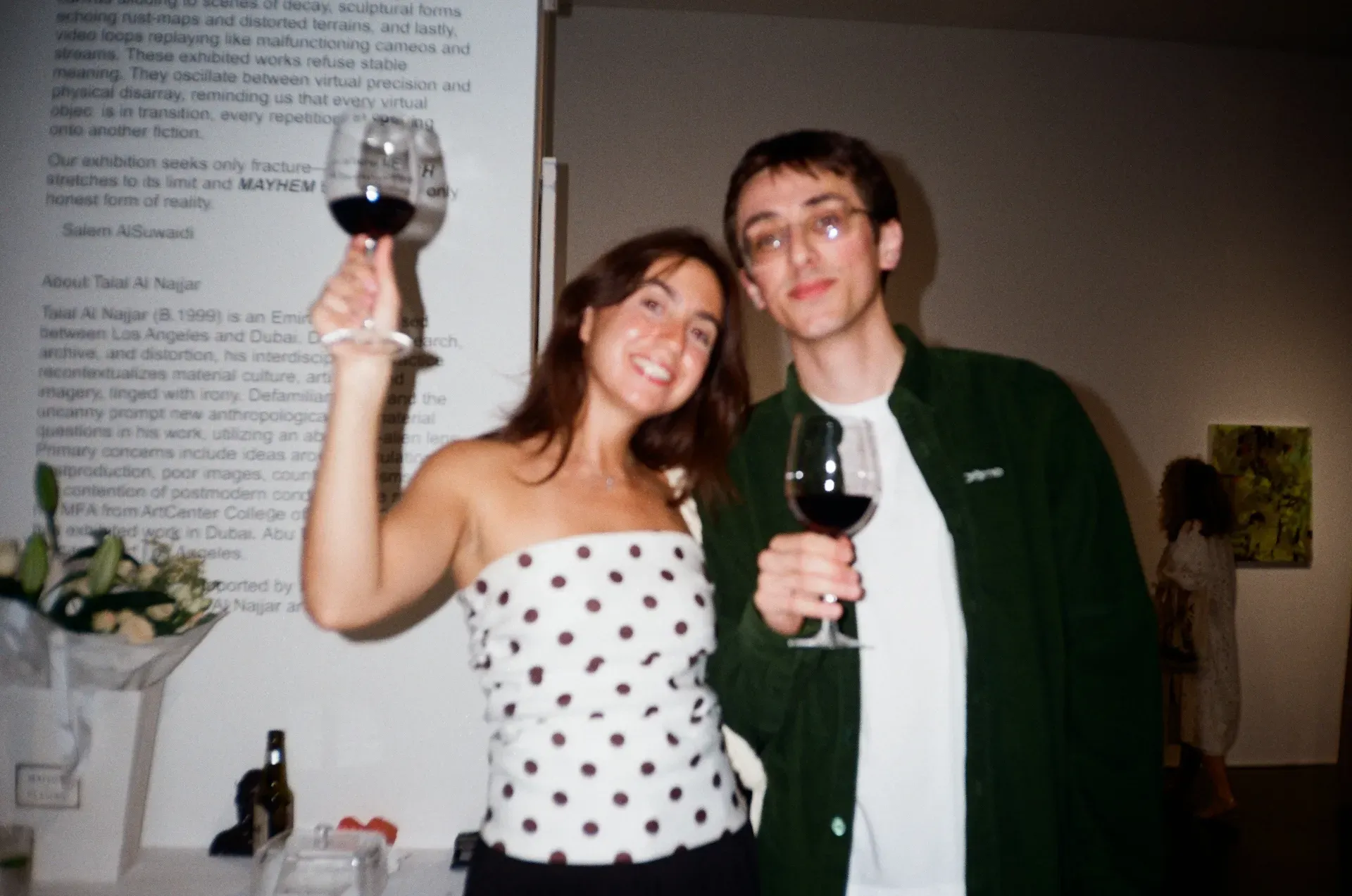 Two guests raising wine glasses in front of the exhibition wall text crediting Salem AlSuwaidi and Talal Al Najjar